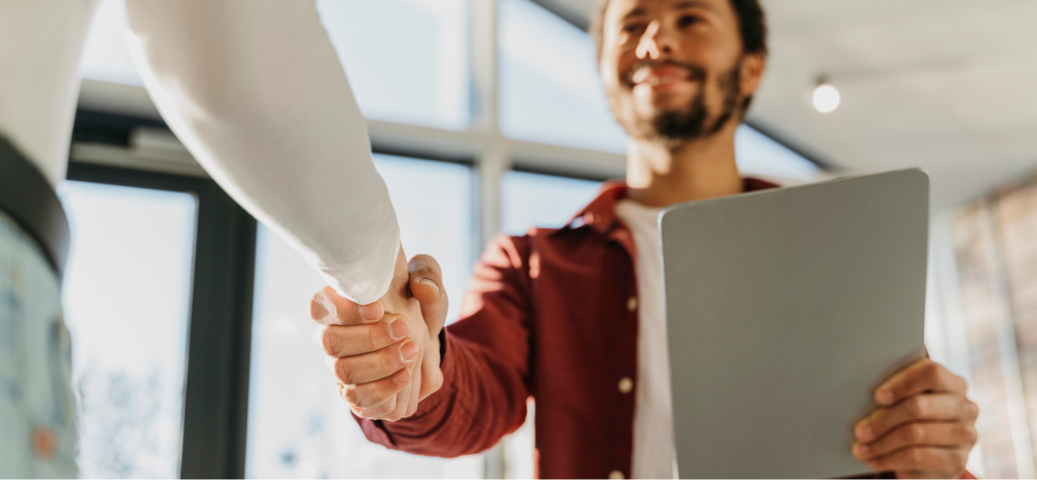 Homem sorrindo segura tablet e aperta a mão de outra pessoa em escritório iluminado pelo sol durante reunião profissional.