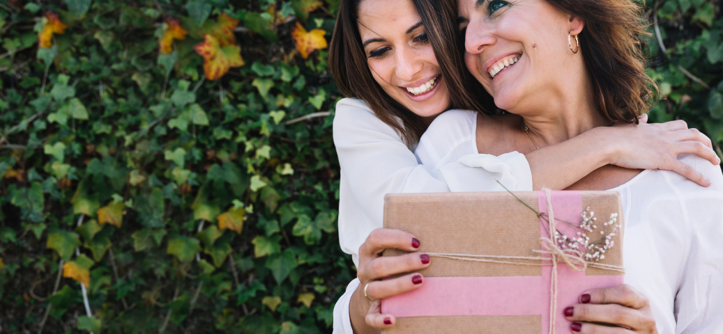 Duas mulheres sorriem e se abraçam enquanto seguram um presente embrulhado em papel pardo com fita rosa e flores secas.