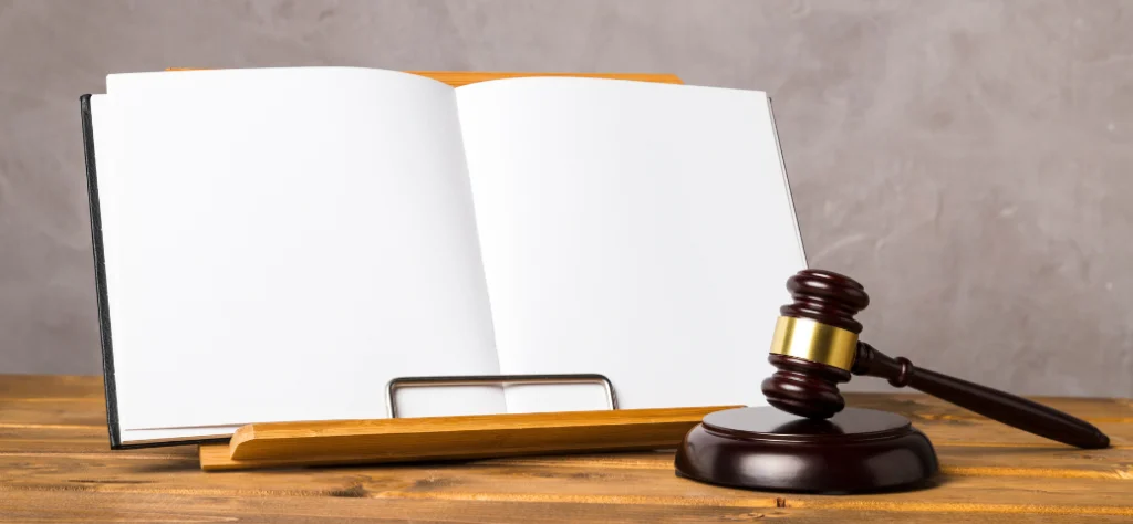 Livro aberto com páginas em branco em um suporte de madeira ao lado de um martelo de juiz (gavel) sobre uma mesa.