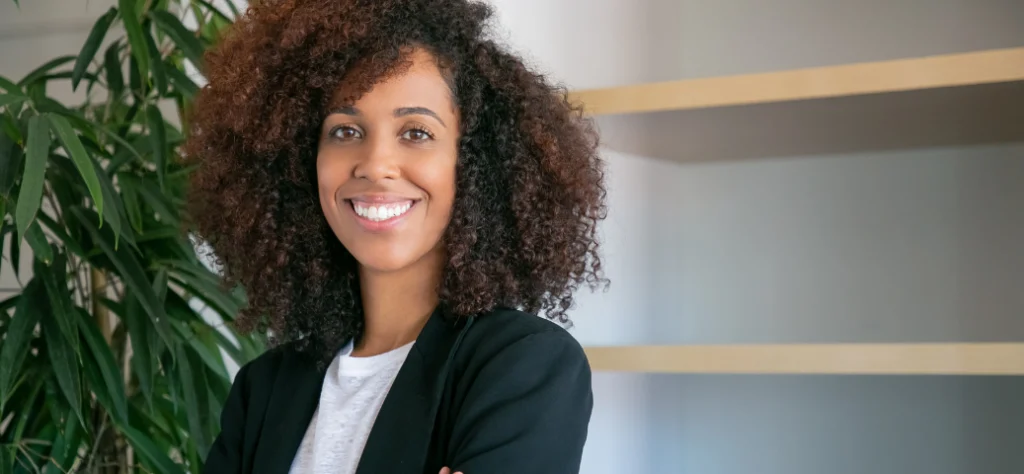 Mulher negra sorridente com cabelo volumoso e blazer escuro posa ao lado de planta e prateleiras em um escritório.