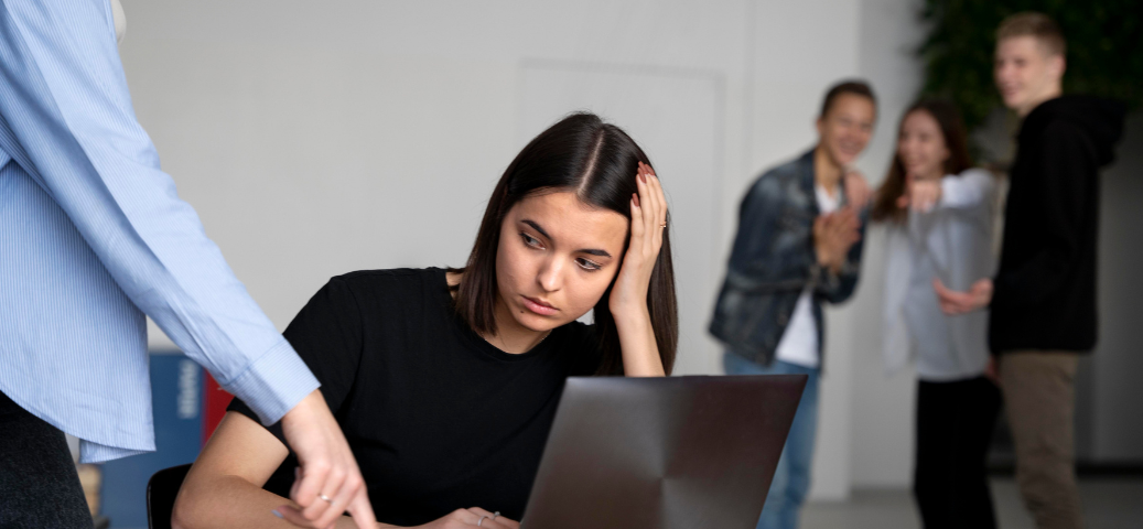 Jovem angustiada no laptop sofre pressão: alguém aponta para a tela enquanto grupo ao fundo ri e faz bullying com ela.
