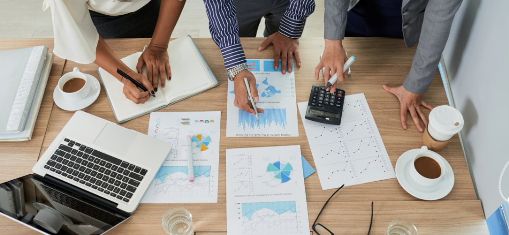 Vista aérea de equipe analisando dados financeiros em gráficos impressos e laptop sobre mesa de madeira, com café e calculadora.
