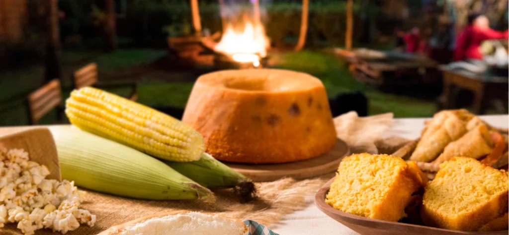 Mesa com comidas típicas de festa junina, como milho, bolo de fubá e pipoca, com uma fogueira acesa ao fundo à noite.