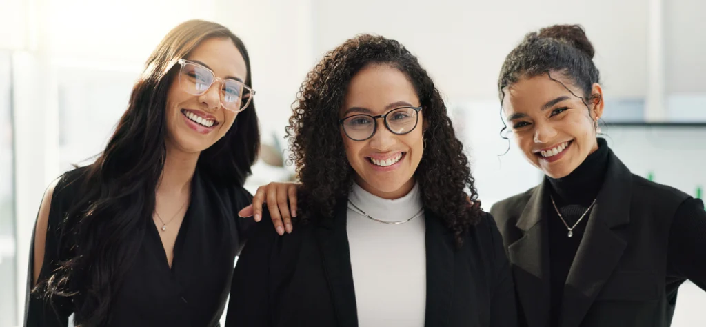 Três mulheres jovens e diversas sorriem juntas para a foto. Elas usam roupas sociais pretas e brancas em um escritório.