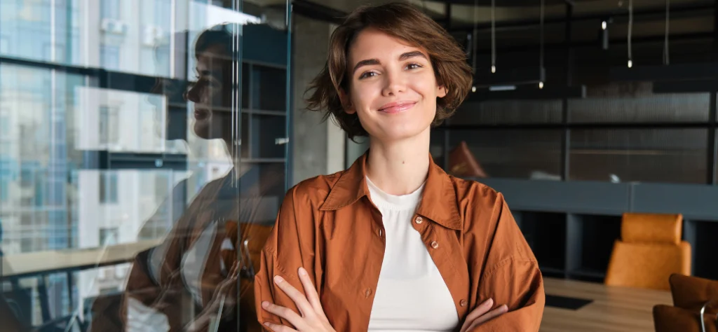 Mulher de cabelo curto e camisa marrom sorri de braços cruzados em escritório, ao lado de um vidro reflexivo.