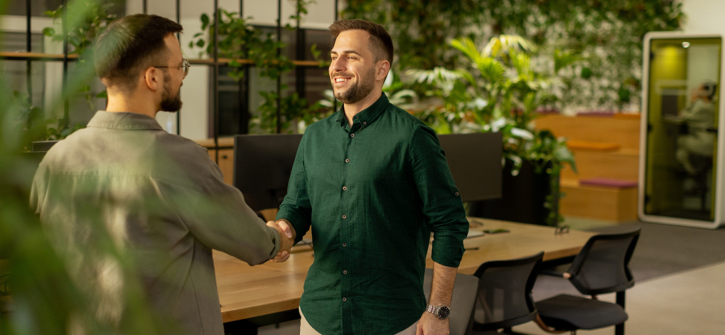 Dois homens sorridentes apertam as mãos em um escritório moderno, com plantas ao fundo