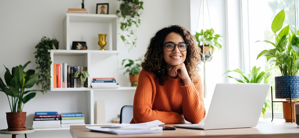 Mulher de óculos e blusa laranja sorri em frente ao notebook, em um escritório decorado com muitas plantas e prateleiras brancas.
