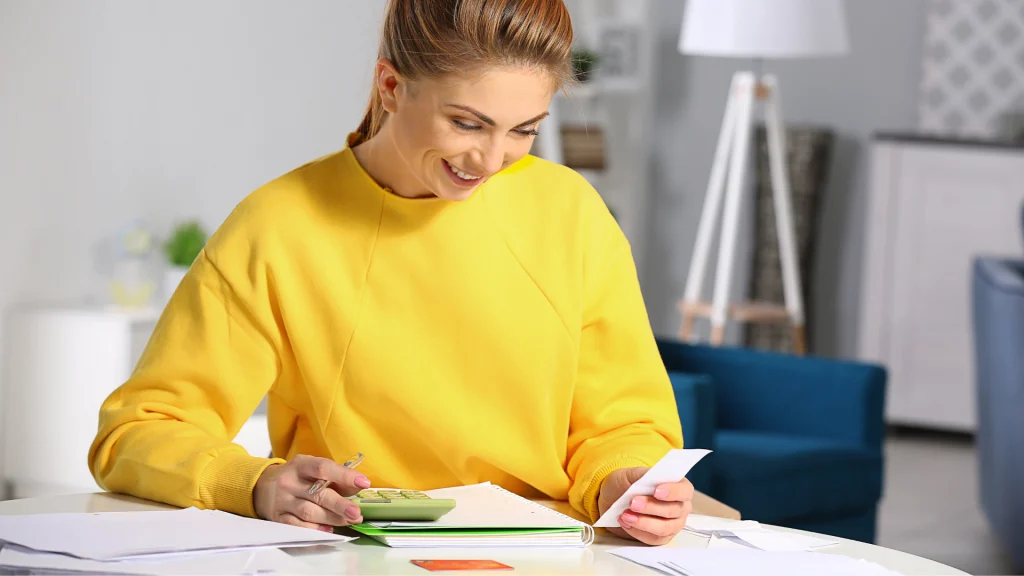 Mulher de moletom amarelo sorri ao usar calculadora e conferir contas em uma mesa com caderno e papéis em casa.