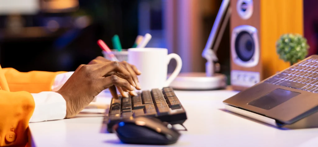 Mãos negras digitam em um teclado em ambiente iluminado à noite, com notebook, caneca e caixa de som ao fundo.