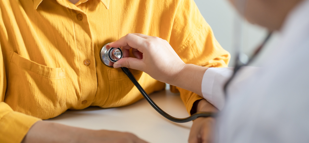 Médico usa estetoscópio para auscultar o coração de paciente de camisa amarela, em consulta médica detalhada.