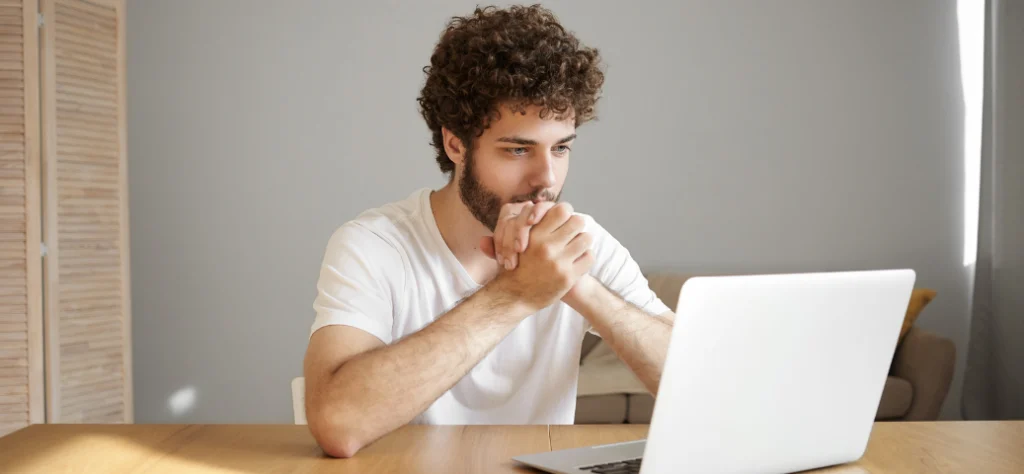 Homem jovem de cabelo cacheado e barba foca a atenção na tela de um notebook, com as mãos entrelaçadas em mesa de madeira.