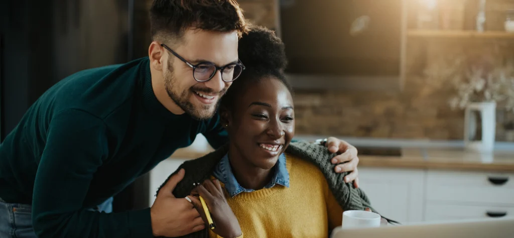 Casal sorridente olha para a tela de um notebook na cozinha, planejando juntos suas metas financeiras com otimismo.