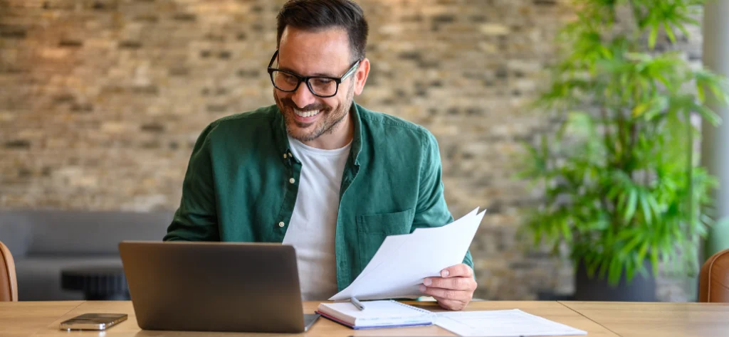 Homem de óculos e camisa verde sorri enquanto analisa documentos em papel e trabalha com notebook em um escritório.