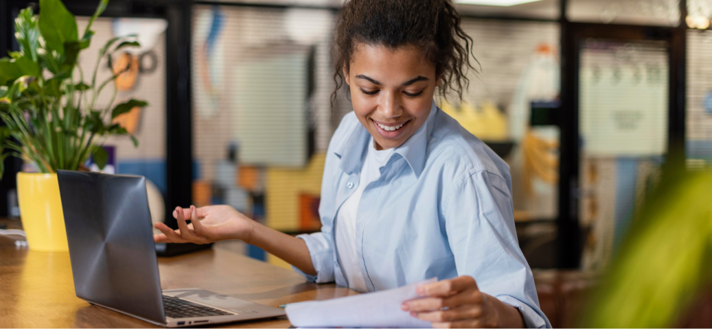 Mulher jovem sorri ao ler um documento em frente ao notebook, com gesto de satisfação em um escritório moderno e iluminado.