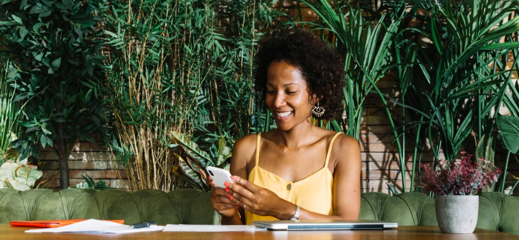 Mulher negra sorridente de blusa amarela olha para o celular, sentada em frente a uma parede de plantas