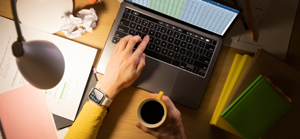 Visão aérea de mãos digitando em um notebook e segurando uma caneca de café em uma mesa com livros, papéis e luminária.