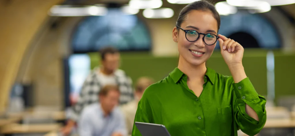 Mulher sorridente de camisa verde e óculos segura um tablet em um escritório, com colegas desfocados ao fundo.