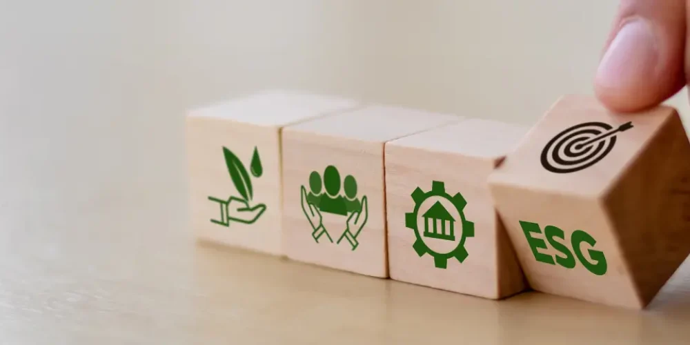 Mão colocando um bloco com "ESG" em uma série de blocos de madeira representando ícones de sustentabilidade, responsabilidade social e governança.