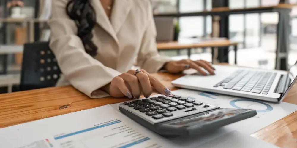 Uma mulher com um blazer claro opera uma calculadora enquanto trabalha em um laptop, com documentos financeiros em uma mesa de madeira em um escritório iluminado.