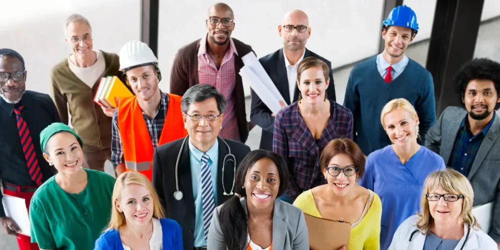Um grupo diversificado de profissionais em trajes variados, incluindo uniformes médicos, capacetes de construção e ternos, reuniu-se em um local fechado.