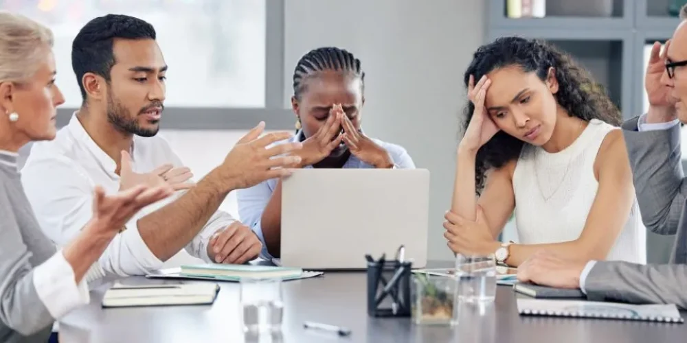 Foto de uma reunião de negócios tensa com cinco pessoas ao redor de uma mesa de conferência. Ao centro, uma mulher negra com tranças cobre o rosto com as mãos, aparentando estar angustiada ou sobrecarregada; um laptop está aberto à sua frente. À sua direita, uma mulher com longos cabelos escuros cacheados apoia a cabeça na mão, com uma expressão de cansaço e frustração. À esquerda da mulher angustiada, um homem de barba e camisa branca gesticula ativamente enquanto fala, parecendo estar em uma discussão acalorada ou tentando se explicar para uma mulher mais velha, de cabelos loiros curtos, que está à sua esquerda e também gesticula enfaticamente. No extremo direito da imagem, um homem mais velho de óculos e terno leva a mão à testa, com uma expressão de preocupação ou exasperação. Sobre a mesa, há cadernos, copos com água e material de escritório. A linguagem corporal de todos sugere um ambiente de conflito, estresse ou desacordo.