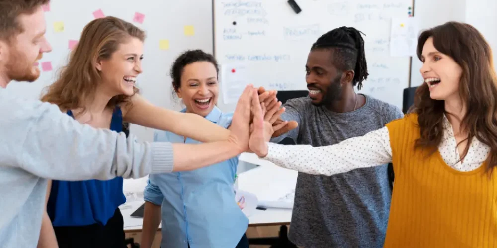A imagem mostra cinco pessoas, três mulheres e dois homens, em um ambiente de escritório ou sala de reunião, participando de um "high five" coletivo, onde suas mãos se encontram no centro em um gesto de celebração ou trabalho em equipe. Todos estão sorrindo e parecem felizes ou entusiasmados.