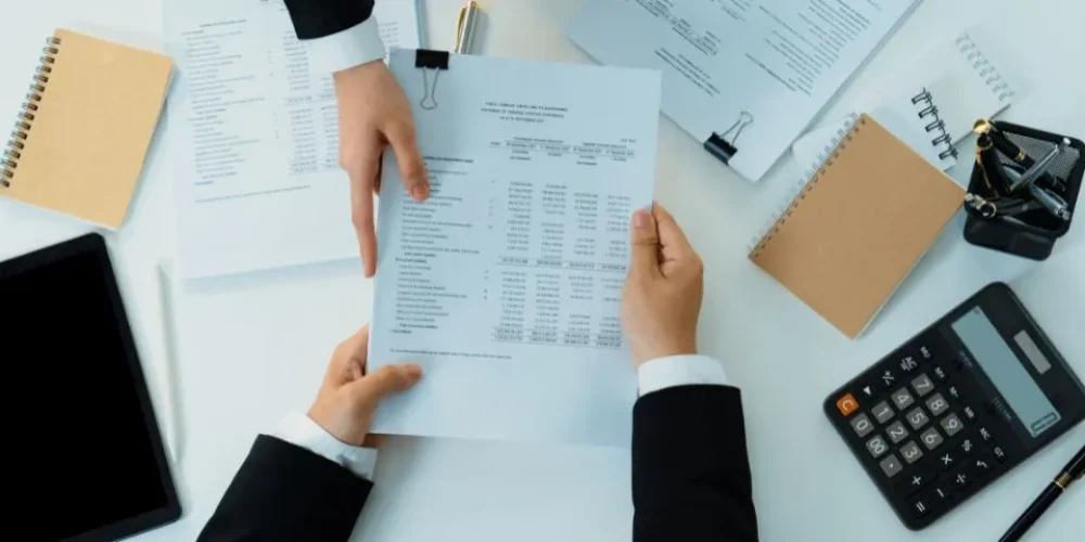 Duas mãos em trajes profissionais trocam um documento financeiro sobre uma mesa desorganizada com uma calculadora, um tablet e blocos de notas.