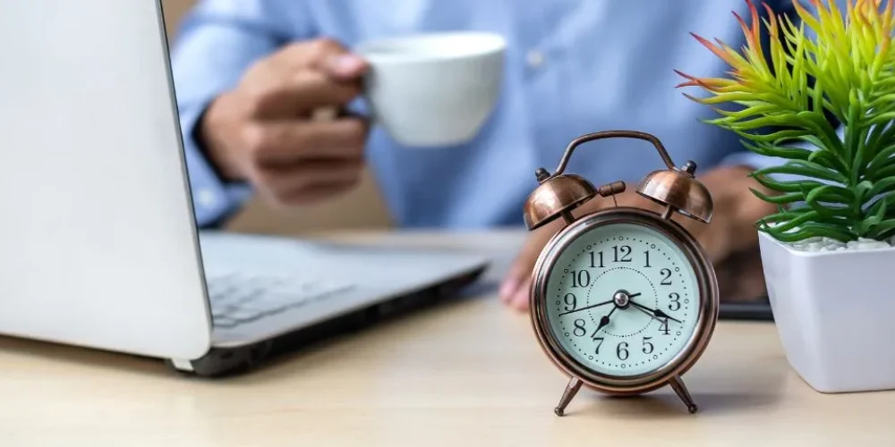 A imagem mostra uma cena em um escritório ou home office, com o foco principal em um despertador analógico de cor cobre, vintage, posicionado à direita no primeiro plano. O despertador tem um mostrador branco com números arábicos pretos e ponteiros pretos, e está obre uma mesa de madeira clara.