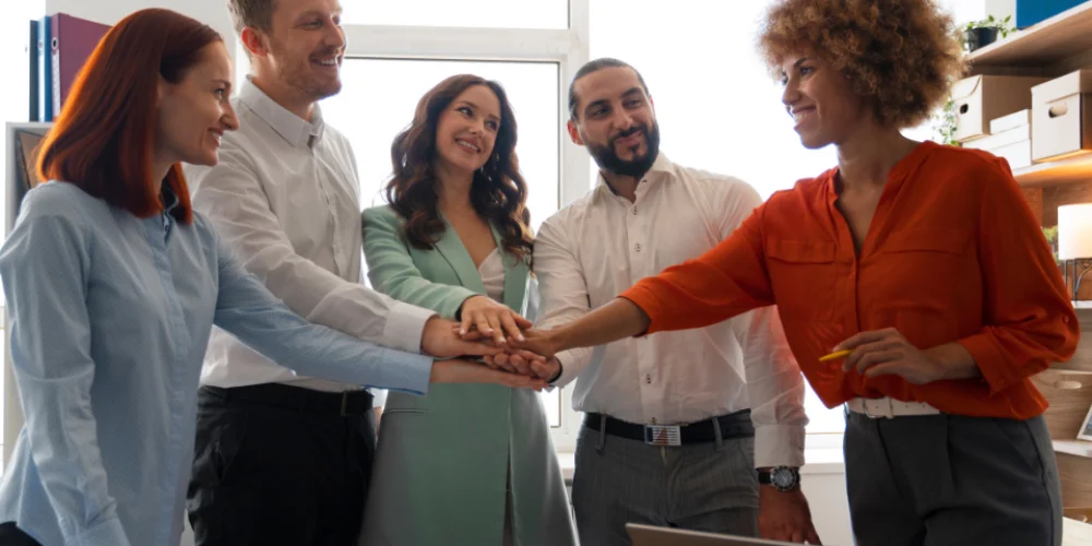 Uma equipe diversa de cinco profissionais, incluindo homens e mulheres, com diferentes etnias e tipos de cabelo, está reunida em um escritório. Eles sorriem e empilham as mãos uma sobre a outra no centro, transmitindo a ideia de união e trabalho em equipe.