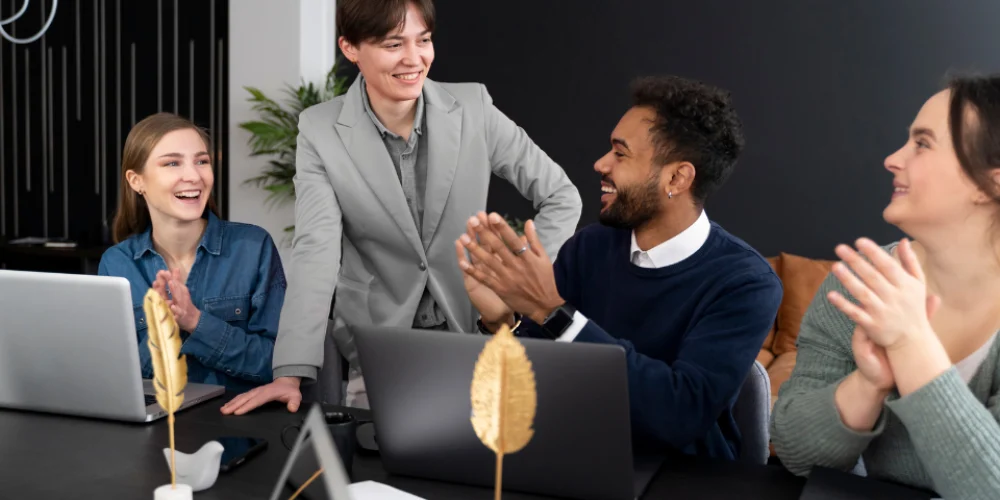 Foto de um ambiente de trabalho positivo. Uma pessoa de blazer cinza está em pé, sorrindo, enquanto três colegas sentados à sua frente a aplaudem com entusiasmo.