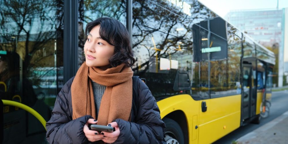 A imagem mostra uma jovem de etnia asiática, com cabelo escuro e curto, em um ambiente urbano. Ela está vestindo um casaco puffer preto sobre um suéter cinza e um cachecol marrom claro bem enrolado no pescoço, o que sugere um clima frio. A jovem está segurando um smartphone com as duas mãos e olhando para cima e para a direita com uma expressão pensativa ou curiosa. Ao fundo, parcialmente visível e ligeiramente desfocado, há um ônibus moderno de cor amarela com detalhes em preto e janelas espelhadas, refletindo árvores e edifícios. A reflexão do ônibus nas janelas parece ter um letreiro digital que, embora ilegível, sugere ser um ônibus de transporte público. O cenário sugere que ela está em uma parada de ônibus ou esperando por um transporte.