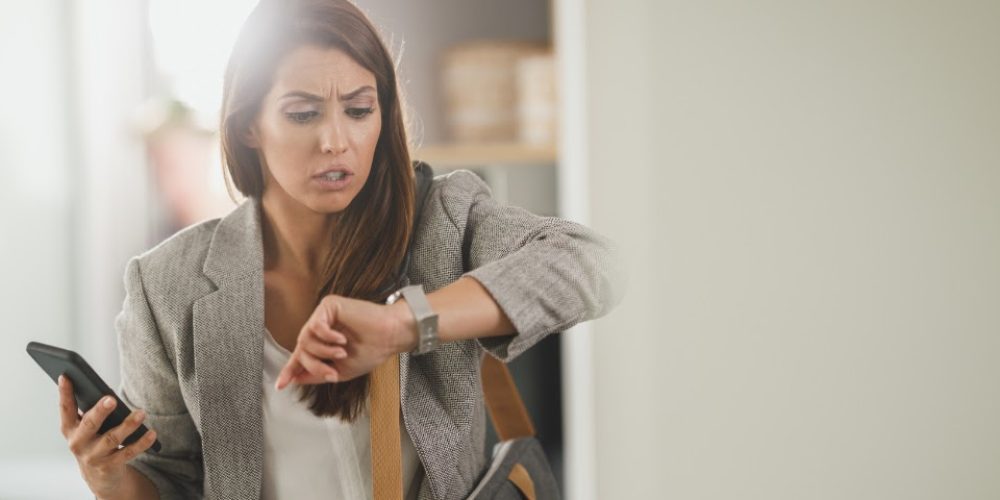 A imagem mostra uma mulher de meia-idade, com cabelos castanhos longos e lisos, vestindo um blazer cinza claro e uma blusa branca. Ela tem uma expressão de preocupação ou surpresa em seu rosto, com as sobrancelhas franzidas e a boca ligeiramente aberta, como se estivesse resmungando ou chocada. Seu olhar está fixo em seu pulso esquerdo, onde ela está olhando um relógio de pulso prateado. Na mão direita, ela segura um smartphone preto. No ombro esquerdo, ela parece carregar uma bolsa escura ou mochila. O fundo está ligeiramente desfocado, mas parece ser um ambiente interno, possivelmente um escritório ou uma casa, com luz natural vindo da esquerda, criando um certo brilho na imagem. A composição sugere que ela está com pressa, atrasada ou surpresa com o horário que vê no relógio.