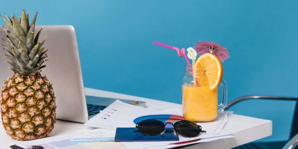 Mesa de escritório com abacaxi, laptop, documentos, óculos de sol e bebida tropical com laranja, canudo e guarda-chuva.