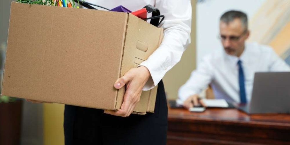 Mulher demitida segura uma caixa de papelão com seus pertences de escritório, enquanto um homem de terno, possivelmente o chefe, está sentado à mesa atrás dela.