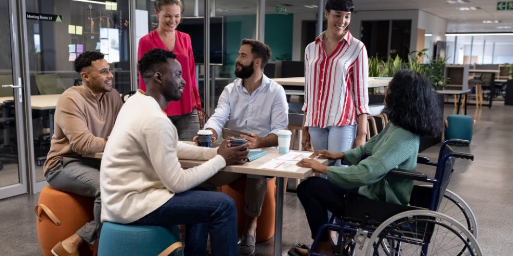 A imagem mostra um grupo diverso de seis adultos em um ambiente de escritório moderno e aberto. Eles estão reunidos em torno de uma pequena mesa de madeira clara, interagindo e parecendo engajados em uma discussão ou reunião informal. Da esquerda para a direita, temos: Um homem de pele escura com um moletom cor de areia e calças cinzas, sentado em um pufe laranja. Ele está sorrindo e olhando para o centro do grupo. Um homem negro de pele escura com uma camisa creme de manga comprida e calça jeans, sentado em um pufe azul-esverdeado. Ele está segurando uma caneca e olhando para o homem à sua frente. Uma mulher branca com uma blusa rosa vibrante e calça escura, que está de pé atrás da mesa e sorrindo para o grupo. Um homem barbudo com pele clara e uma camisa social branca de manga comprida, sentado na mesa e segurando uma caneca. Ele está olhando para a mulher de camisa rosa. Uma mulher com pele clara e cabelo escuro, usando uma camisa listrada vermelha e branca e calça jeans. Ela está de pé do outro lado da mesa, olhando para o centro do grupo. Uma mulher negra sentada em uma cadeira de rodas, usando um suéter verde escuro e calças escuras. Ela está virada para a mesa e parece estar participando ativamente da conversa.