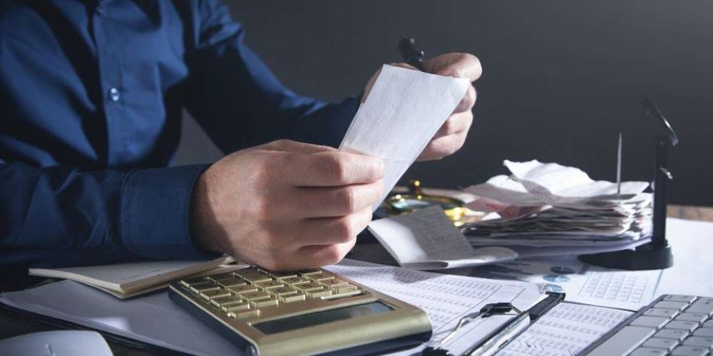 Mãos de uma pessoa segurando um pequeno pedaço de papel branco, possivelmente um recibo. Ao fundo, uma mesa bagunçada com uma pilha de recibos e uma calculadora dourada.