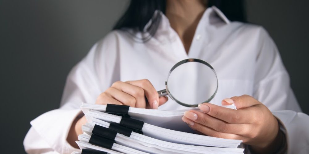 A imagem mostra uma pessoa segurando uma lupa sobre uma pilha de documentos. A pessoa está vestindo uma camisa branca e tem unhas bem cuidadas. A pilha de documentos é grossa e está presa com vários clipes pretos, indicando que são muitos papéis. A lupa está posicionada no centro da imagem, com as mãos da pessoa segurando-a logo abaixo. O fundo é escuro e desfocado, o que faz com que o foco principal da imagem esteja na lupa e nos documentos. A iluminação é suave, destacando os objetos em primeiro plano. A imagem sugere um tema de pesquisa, análise, auditoria ou verificação de informações.