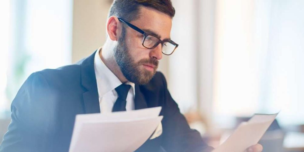 Um homem barbudo de terno e óculos lendo papéis em um escritório.