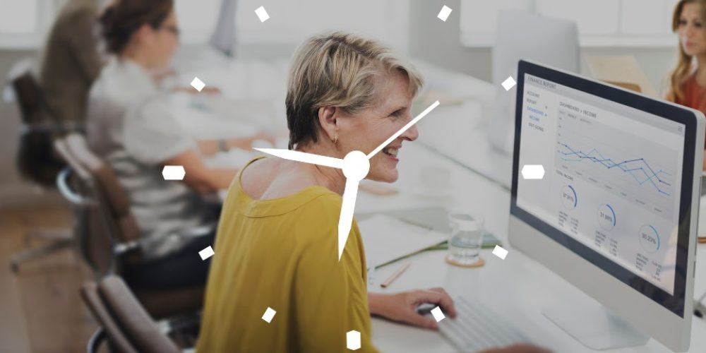 A imagem mostra um ambiente de escritório com uma mulher de meia-idade, com cabelos loiros curtos, sorrindo enquanto olha para um monitor de computador à sua frente. Ela veste uma blusa amarela. À esquerda da mulher, há outras pessoas desfocadas trabalhando em suas mesas, e à direita, outra pessoa desfocada pode ser vista ao fundo. A tela do computador exibe gráficos e dados, provavelmente relacionados a finanças ou relatórios. Sobreposta à imagem, há uma representação de um relógio analógico branco, com ponteiros que indicam aproximadamente 10 horas e 10 minutos (o ponteiro das horas está um pouco antes do 2 e o dos minutos está no 2).