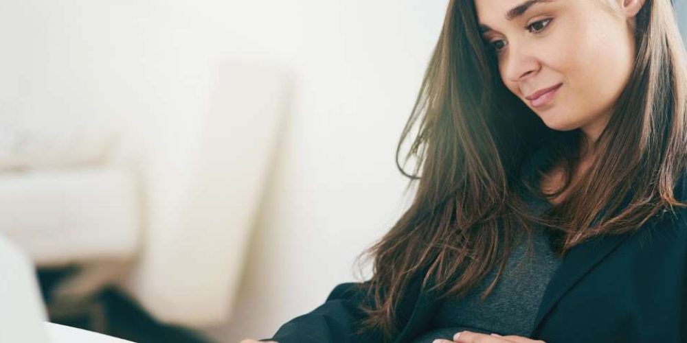 Uma mulher grávida com cabelo castanho escuro e uma blusa cinza, vestindo um blazer preto, está sentada em uma mesa e olha para baixo com uma expressão calma e gentil. Ela apoia a mão na barriga e parece estar tocando na tela de um notebook que não aparece na imagem.