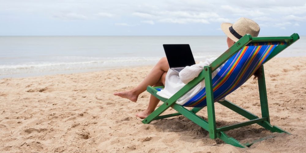 A imagem mostra uma pessoa sentada em uma cadeira de praia listrada, de costas para o observador, com vista para o oceano. A pessoa está usando um chapéu de palha e uma roupa clara, e parece estar utilizando um laptop que está apoiado em seu colo. A cena se passa em uma praia, com areia clara em primeiro plano e as ondas do mar suaves ao fundo. O céu está parcialmente nublado, e a linha do horizonte entre o mar e o céu é visível. A cadeira tem uma estrutura de madeira verde e um tecido com listras coloridas em azul, branco, amarelo, laranja e verde. A impressão geral é de relaxamento e trabalho remoto em um ambiente de férias.