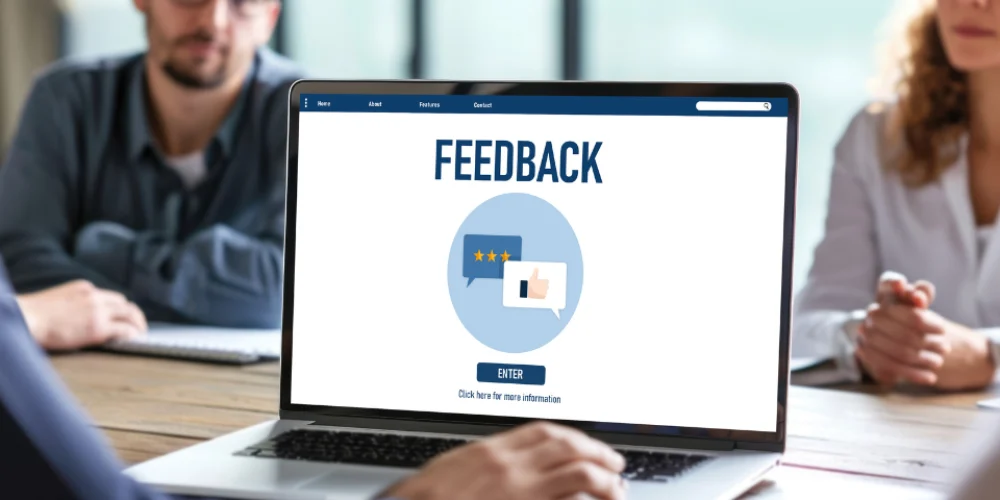 Uma visão aproximada de um laptop em uma mesa de conferência, com a palavra "FEEDBACK" exibida na tela. Abaixo da palavra, há dois balões de fala em um círculo azul claro, um com estrelas de avaliação e outro com um ícone de polegar para cima. O fundo mostra pessoas borradas sentadas ao redor da mesa.